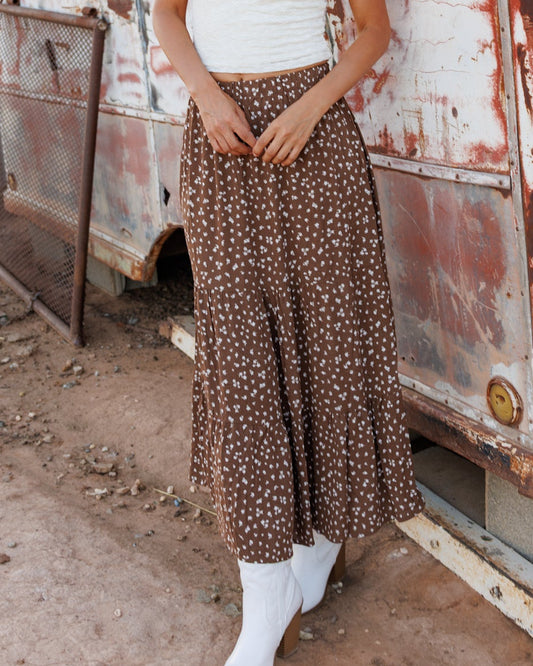 A woman stands outdoors in front of a weathered, rusted trailer wearing a white short-sleeve top, a brown floral maxi skirt, a black cowboy hat, and white cowboy boots.