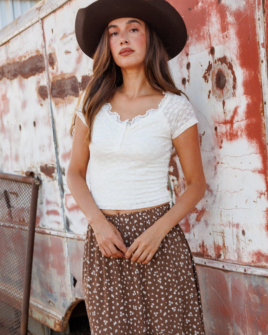 A woman stands outdoors in front of a weathered, rusted trailer wearing a white short-sleeve top, a brown floral maxi skirt, a black cowboy hat, and white cowboy boots.