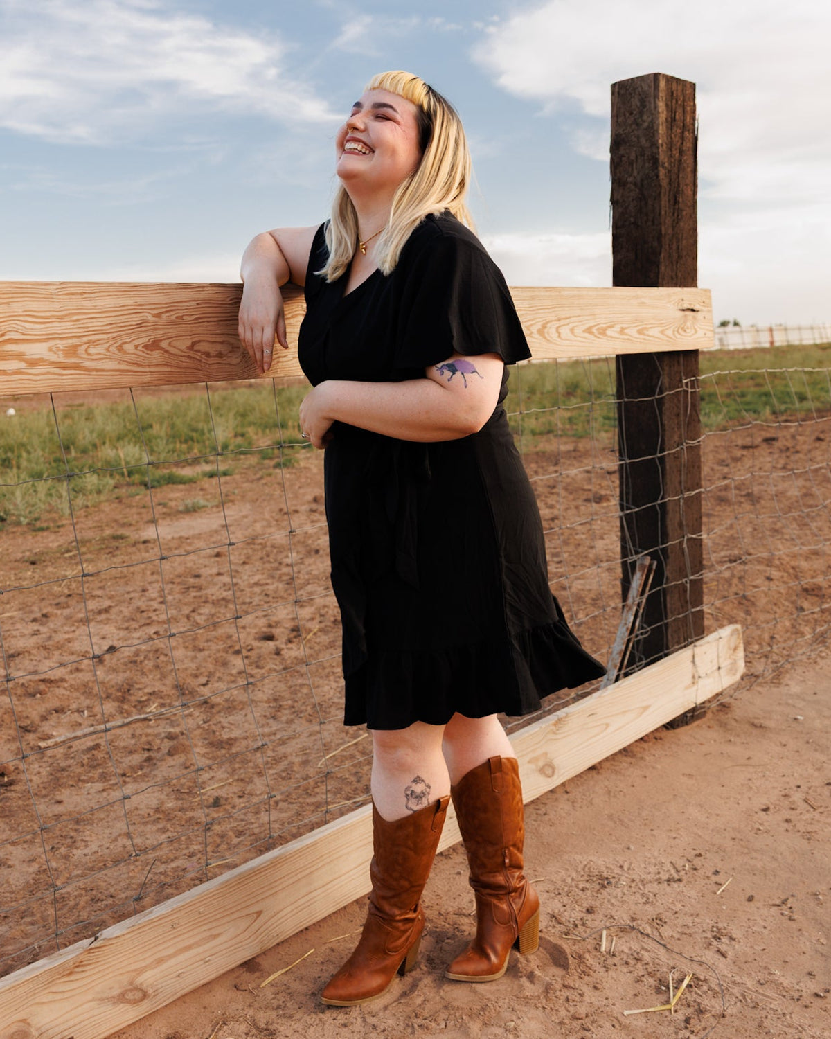 A woman with blonde hair wearing a black dress and brown cowboy boots leans against a wooden fence in an outdoor field under a blue sky.