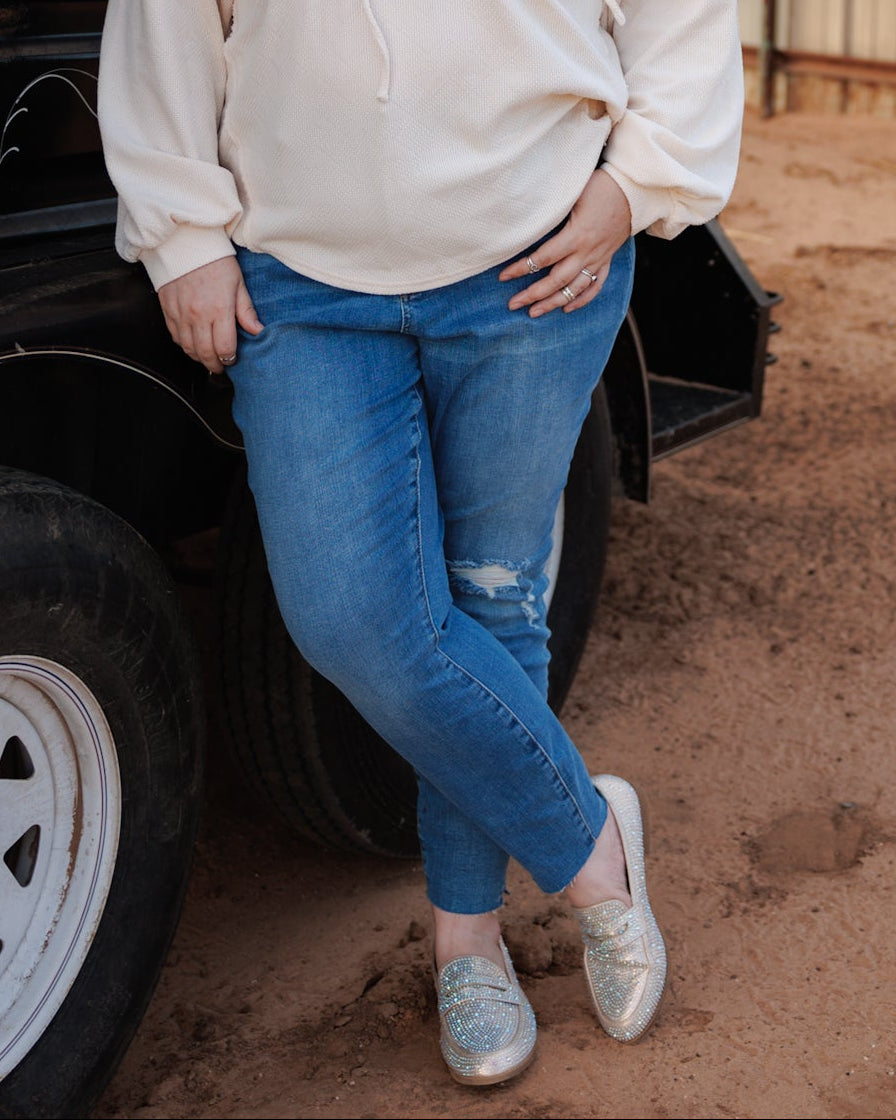 A woman stands outdoors next to a black livestock trailer on a dirt ground, wearing a cream-colored long-sleeve hoodie top, blue jeans, and rhinestone beige loafers.