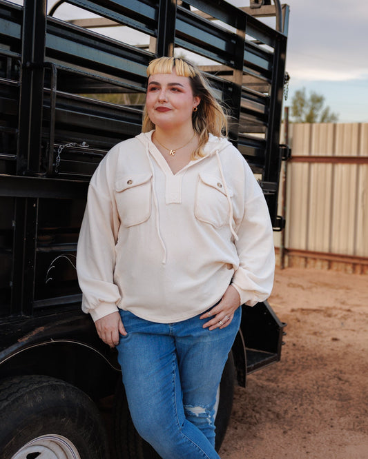 A woman stands outdoors next to a black livestock trailer on a dirt ground, wearing a cream-colored long-sleeve button-up shirt, blue jeans, and silver sneakers.
