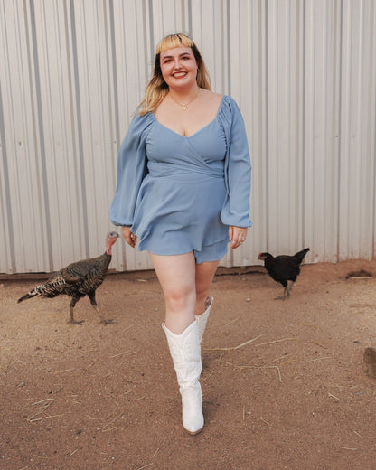 A woman stands outdoors in front of a corrugated metal wall, wearing a light blue wrap-style mini romper dress with long puff sleeves and white cowboy boots. Two chickens and a turkey are visible on the dirt ground nearby.